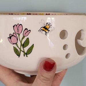 Op de afbeelding: Crèmekleurige keramische garenkom met een bloemen- en bijenontwerp. De kom heeft roze tulpen en een cartoonbij. Het heeft een gebogen sleuf en verschillende gaten voor garenbeheer.