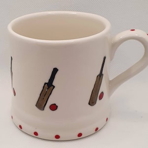 May include: White ceramic mug with a red and brown cricket bat design. The mug has a red polka dot pattern around the bottom.