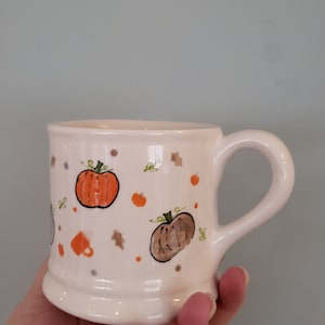 Pode incluir: Caneca de cerâmica creme decorada com abóboras laranja e marrons, folhas e pontos. A caneca tem uma alça arredondada e uma base ligeiramente alargada. Um design com tema de outono.