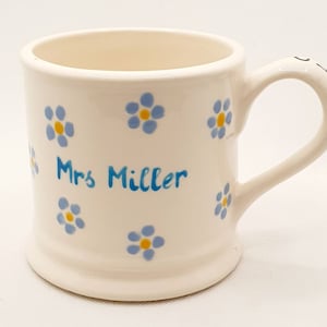 Puede incluir: Taza de cerámica color crema decorada con motivos florales azules y amarillos. La taza tiene un asa curvada y el nombre "Mrs Miller" escrito en azul. Un objeto encantador para té o café, ideal para el uso diario.