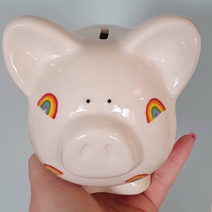 Puede incluir: Huchita de cer&aacute;mica blanca con calcoman&iacute;as de arco&iacute;ris. La alcanc&iacute;a tiene una ranura en la parte superior para monedas.
