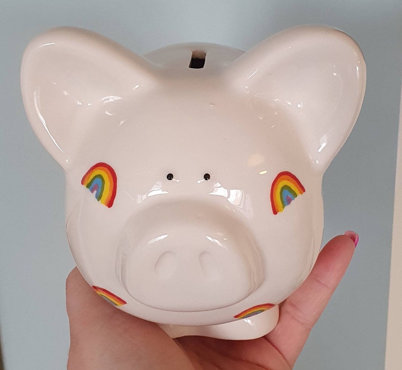 Puede incluir: Huchita de cer&aacute;mica blanca con calcoman&iacute;as de arco&iacute;ris. La alcanc&iacute;a tiene una ranura en la parte superior para monedas.