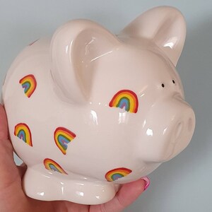 Puede incluir: Una alcanc&iacute;a de cer&aacute;mica blanca con un dise&ntilde;o de arco&iacute;ris. La alcanc&iacute;a tiene un hocico rosa y est&aacute; decorada con varios patrones de arco&iacute;ris.