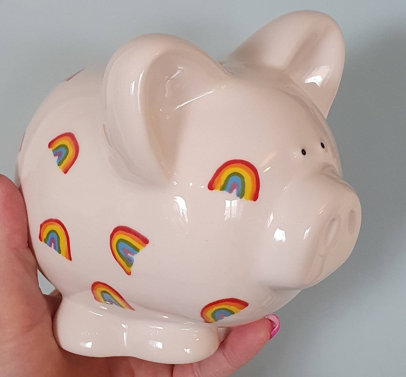 Puede incluir: Una alcanc&iacute;a de cer&aacute;mica blanca con un dise&ntilde;o de arco&iacute;ris. La alcanc&iacute;a tiene un hocico rosa y est&aacute; decorada con varios patrones de arco&iacute;ris.