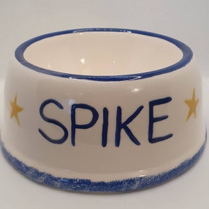May include: A cream-colored ceramic pet bowl with a blue rim and a blue base. The word SPIKE is painted in blue letters on the front, flanked by two yellow stars. The interior of the bowl is smooth and cream-colored, matching the exterior finish.