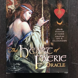 May include: A boxed oracle deck titled "The Heart of Faerie Oracle" by Brian & Wendy Froud. The cover art depicts a woman with red hair and a white gown, holding a staff. The deck's title is in gold lettering, with additional text.