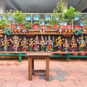 May include: A colorful wooden carving depicting a row of Hindu deities, with intricate details and vibrant colors. The carving is mounted on a wall and surrounded by potted plants. A wooden stool sits in front of the carving.