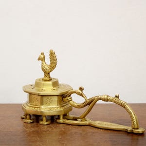 May include: A vintage brass incense burner with a rooster finial. The burner has an octagonal base with decorative cutouts and a handle. The piece is made of brass and sits on a wooden surface.