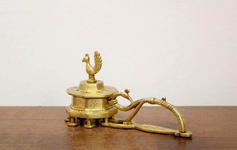 May include: A vintage brass incense burner with a rooster finial. The burner has an octagonal base with decorative cutouts and a handle. The piece is made of brass and sits on a wooden surface.