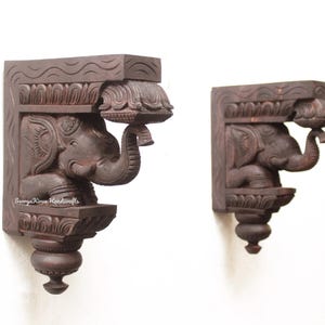 May include: Two dark brown wooden wall brackets with carved elephant figures. The brackets are intricately detailed and have a traditional Indian design.