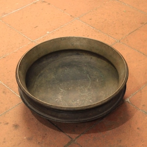 May include: A round, antique metal bowl with a dark interior and a wide, slightly flared rim. The bowl has a dark, aged appearance and sits on a terracotta tile surface. The bowl's design suggests it may be used for decorative purposes.