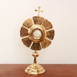 May include: A gold-colored monstrance with a round, ornate base and a central, circular, clear glass container. The monstrance is topped with a cross and has a decorative, flower-shaped design.