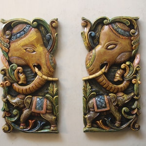 May include: Two intricately carved wooden wall hangings depicting elephants. The elephants are painted in shades of gold, green, blue, and red. The carvings feature detailed floral patterns and a smaller elephant beside each larger elephant.