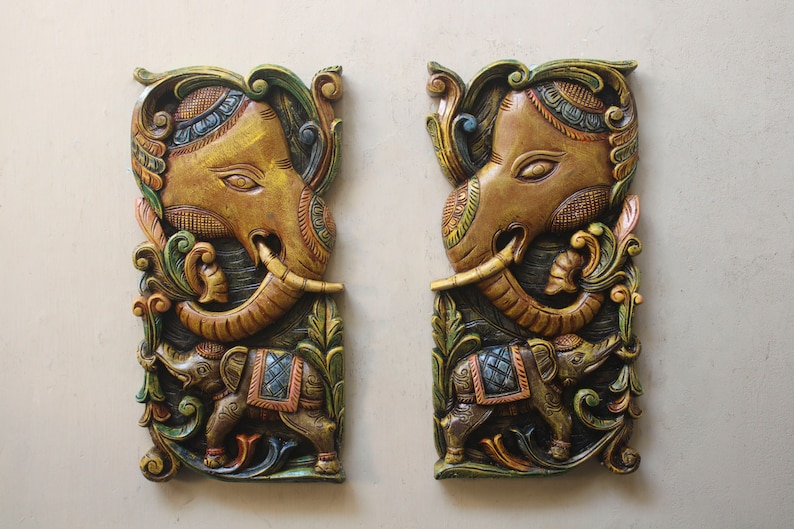 May include: Two intricately carved wooden wall hangings depicting elephants. The elephants are painted in shades of gold, green, blue, and red. The carvings feature detailed floral patterns and a smaller elephant beside each larger elephant.
