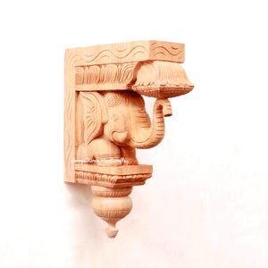 May include: A wooden carved elephant bracket with intricate details. The elephant is holding a decorative lamp. The bracket is a light brown color.