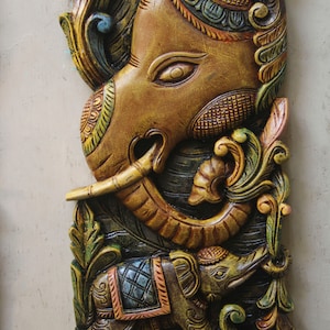 May include: A carved wooden wall hanging featuring two elephants. The larger elephant is brown with intricate details and a raised trunk. The smaller elephant is green and brown with a blue and orange saddle. Both elephants are surrounded by green and gold floral designs.