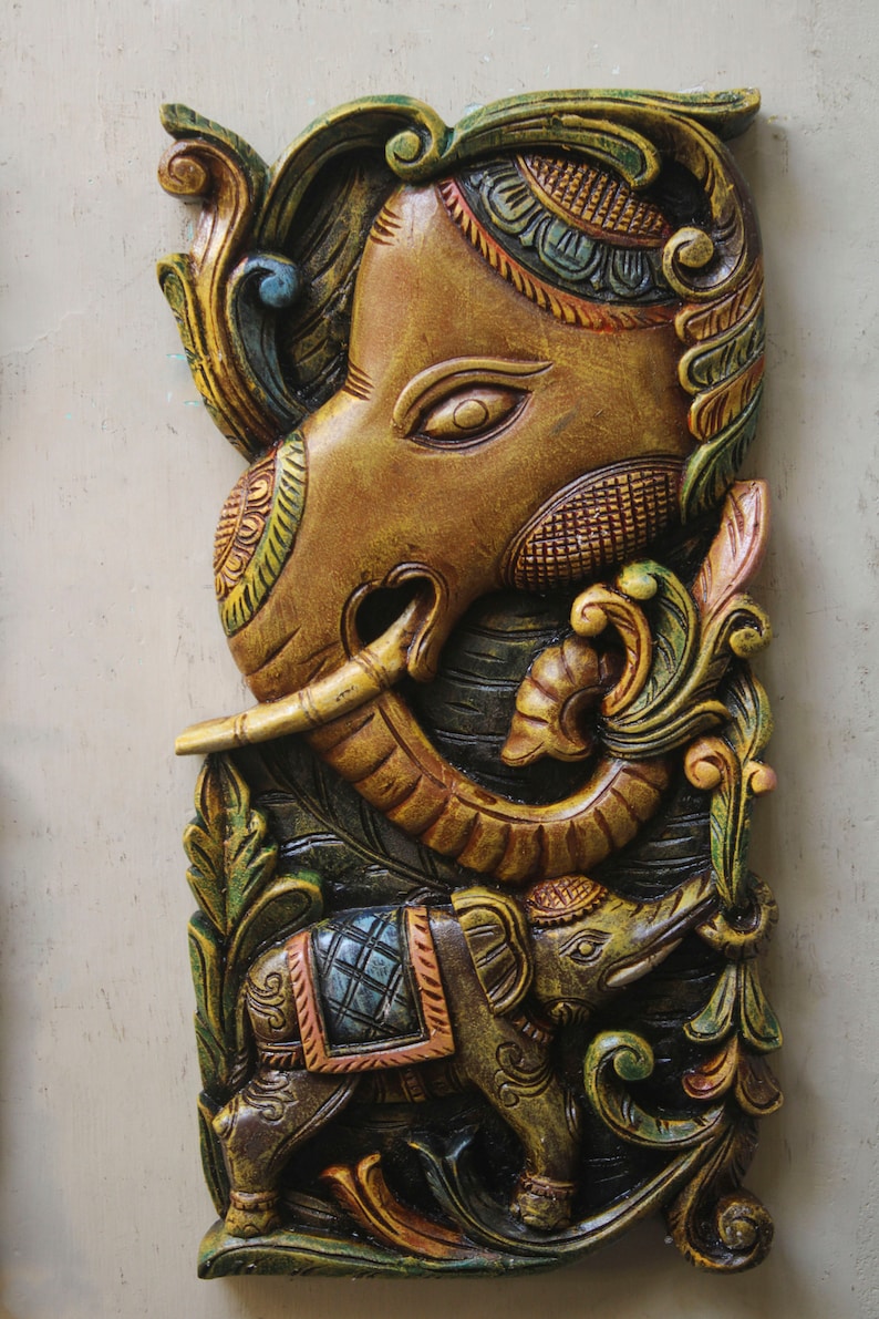 May include: A carved wooden wall hanging featuring two elephants. The larger elephant is brown with intricate details and a raised trunk. The smaller elephant is green and brown with a blue and orange saddle. Both elephants are surrounded by green and gold floral designs.
