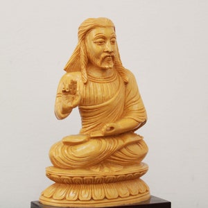 May include: A wooden sculpture of a seated figure with long hair and a beard. The figure is wearing a robe and has a serene expression. The sculpture is carved in a light brown color and is sitting on a base.