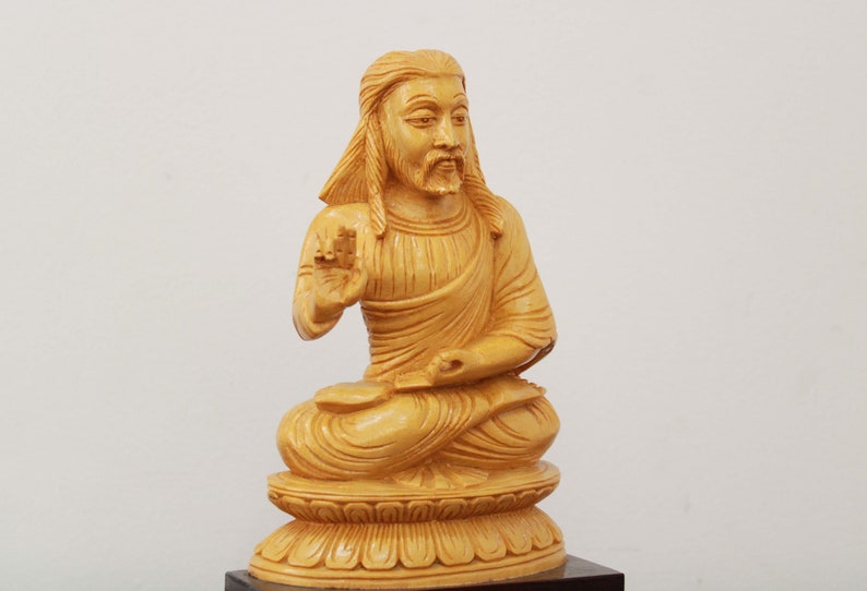 May include: A wooden sculpture of a seated figure with long hair and a beard. The figure is wearing a robe and has a serene expression. The sculpture is carved in a light brown color and is sitting on a base.