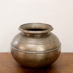 May include: A vintage metal pot with a rounded body and a wide, slightly flared rim. The pot has horizontal bands around its midsection and a tarnished, aged appearance. It sits on a wooden surface against a white background.