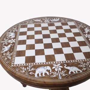 May include: A round wooden chessboard with a white and brown checkered pattern. The board is decorated with a border of white elephants and palm trees.