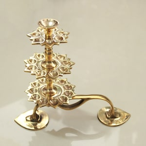 May include: A brass diya lamp with three tiers of petal-shaped holders for oil lamps. The lamp has a curved handle and two leaf-shaped bases. The overall design is ornate and traditional.