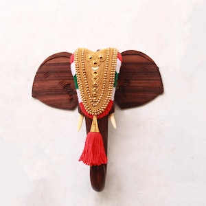 May include: A wooden elephant head wall decoration with intricate carvings. The elephant is adorned with a gold and red garland with a tassel.