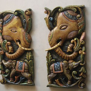 May include: Two intricately carved wooden wall hangings featuring elephant motifs. The elephants are painted in shades of gold, brown, green, and blue, with intricate details and floral patterns.