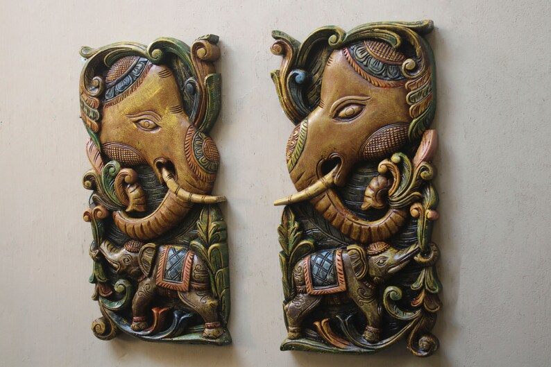 May include: Two intricately carved wooden wall hangings featuring elephant motifs. The elephants are painted in shades of gold, brown, green, and blue, with intricate details and floral patterns.