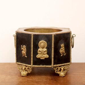 May include: A black and gold hexagonal decorative container with a central Buddha figure and two smaller figures. The container has ornate gold accents and handles, and sits on decorative gold feet.