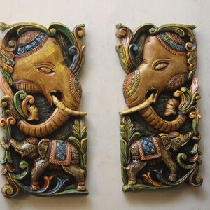 May include: Two intricately carved wooden wall hangings depicting elephants with floral designs. The elephants are painted in shades of brown, gold, green, and blue.