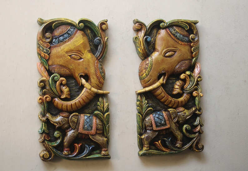 May include: Two intricately carved wooden wall hangings depicting elephants with floral designs. The elephants are painted in shades of brown, gold, green, and blue.