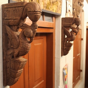 May include: Two intricately carved wooden brackets in the shape of birds with outstretched wings. The brackets are mounted on a white wall and are a traditional architectural element.
