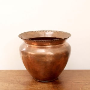 May include: A copper-colored vase with a wide, ruffled rim. The vase has a rounded body and sits on a wooden surface. The interior of the vase is dark. The background is a plain, off-white wall.