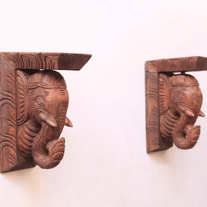 May include: Two intricately carved wooden elephant head wall shelves. The shelves are a warm brown color and feature detailed carvings of elephant heads with trunks and tusks. The shelves are mounted on a white wall.