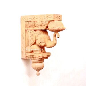 May include: A wooden carved elephant bracket with intricate details. The elephant is holding a small bowl in its trunk. The bracket is a light brown color and has a traditional Indian design.