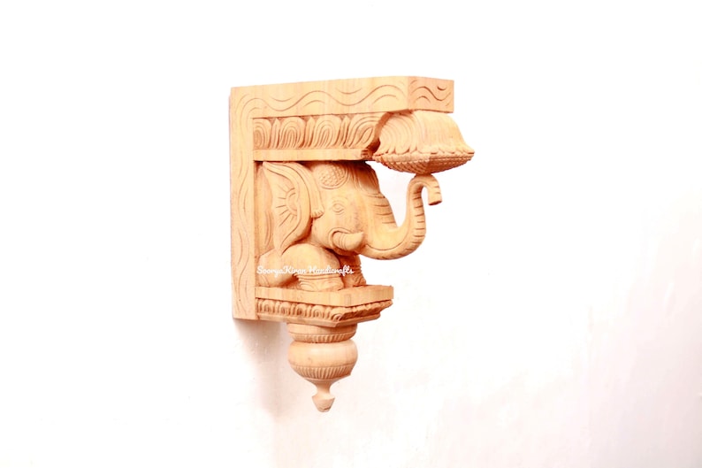 May include: A wooden carved elephant bracket with intricate details. The elephant is holding a small bowl in its trunk. The bracket is a light brown color and has a traditional Indian design.