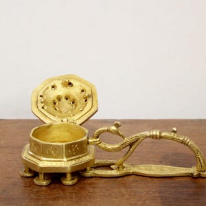 May include: A vintage brass incense burner with an open, octagonal lid and a decorative handle. The burner is gold-colored and sits on small feet. The lid has several small holes.