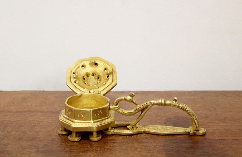 May include: A vintage brass incense burner with an open, octagonal lid and a decorative handle. The burner is gold-colored and sits on small feet. The lid has several small holes.