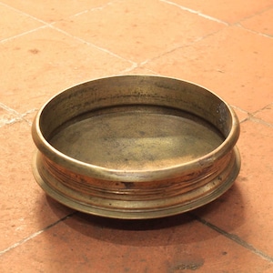 May include: A round, antique brass bowl with a wide rim and a slightly concave interior. The bowl has a warm, golden hue and is set against a terracotta tile background. The bowl's design features a series of concentric rings around its base.