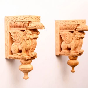 Puede incluir: Dos soportes de pared de madera con intrincados tallados de criaturas míticas aladas. Los soportes son de color marrón claro y tienen un diseño decorativo.