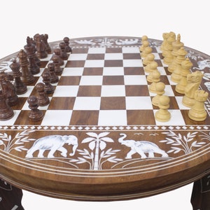 May include: A round wooden chessboard table with intricate carvings of elephants and palm trees. The chessboard is made of dark and light wood squares and features a set of white and brown chess pieces.