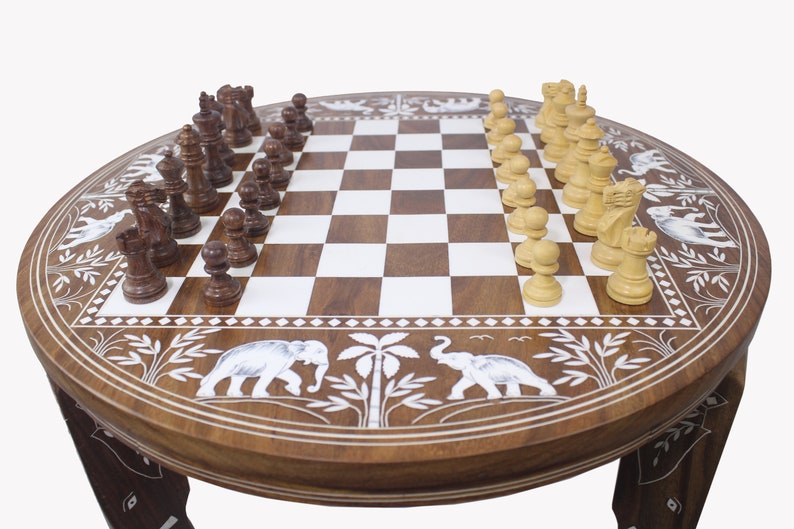 May include: A round wooden chessboard table with intricate carvings of elephants and palm trees. The chessboard is made of dark and light wood squares and features a set of white and brown chess pieces.