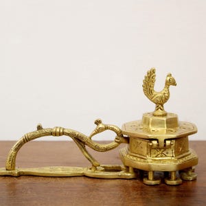 May include: A vintage brass incense burner with a peacock design. The burner features a hinged compartment, a decorative handle, and a peacock figure on top. The brass has a warm, golden hue, and the piece is set on a wooden surface.