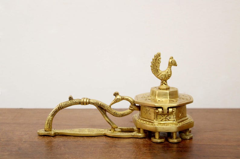 May include: A vintage brass incense burner with a peacock design. The burner features a hinged compartment, a decorative handle, and a peacock figure on top. The brass has a warm, golden hue, and the piece is set on a wooden surface.
