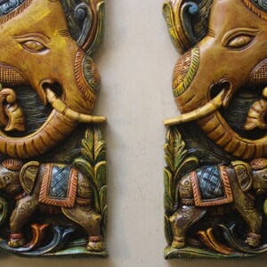 May include: Two intricately carved wooden panels depicting elephants. The panels are painted in shades of gold, brown, green, and blue. Each panel features a larger elephant with its trunk raised, and a smaller elephant standing below. The panels are a traditional Indian art form.
