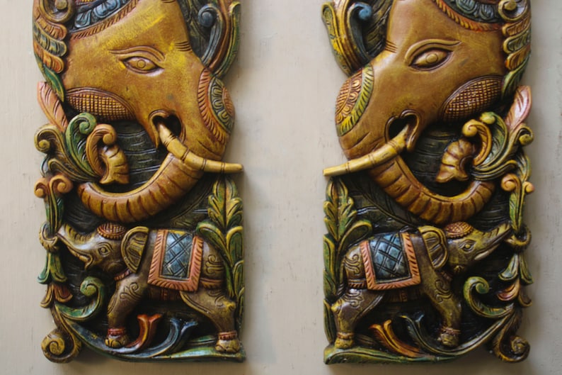 May include: Two intricately carved wooden panels depicting elephants. The panels are painted in shades of gold, brown, green, and blue. Each panel features a larger elephant with its trunk raised, and a smaller elephant standing below. The panels are a traditional Indian art form.