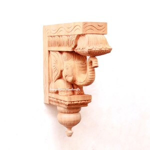May include: A wooden carved elephant bracket with intricate details. The bracket is made of light brown wood and features a detailed carving of an elephant with its trunk raised. The bracket is a traditional Indian design and would be a beautiful addition to any home.