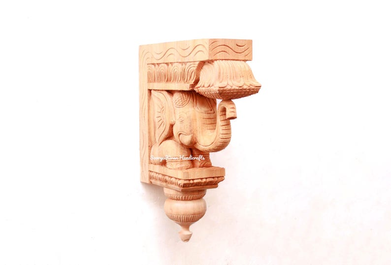 May include: A wooden carved elephant bracket with intricate details. The bracket is made of light brown wood and features a detailed carving of an elephant with its trunk raised. The bracket is a traditional Indian design and would be a beautiful addition to any home.