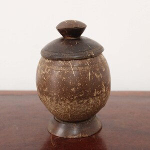 以下が含まれることがあります： 蓋付きの小さな丸い茶色のココナッツの殻の容器。容器は自然で素朴な外観で、木製の表面の上に置かれています